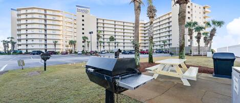 A parking lot with a grill, picnic table, trash bin, and a hotel building in the background.