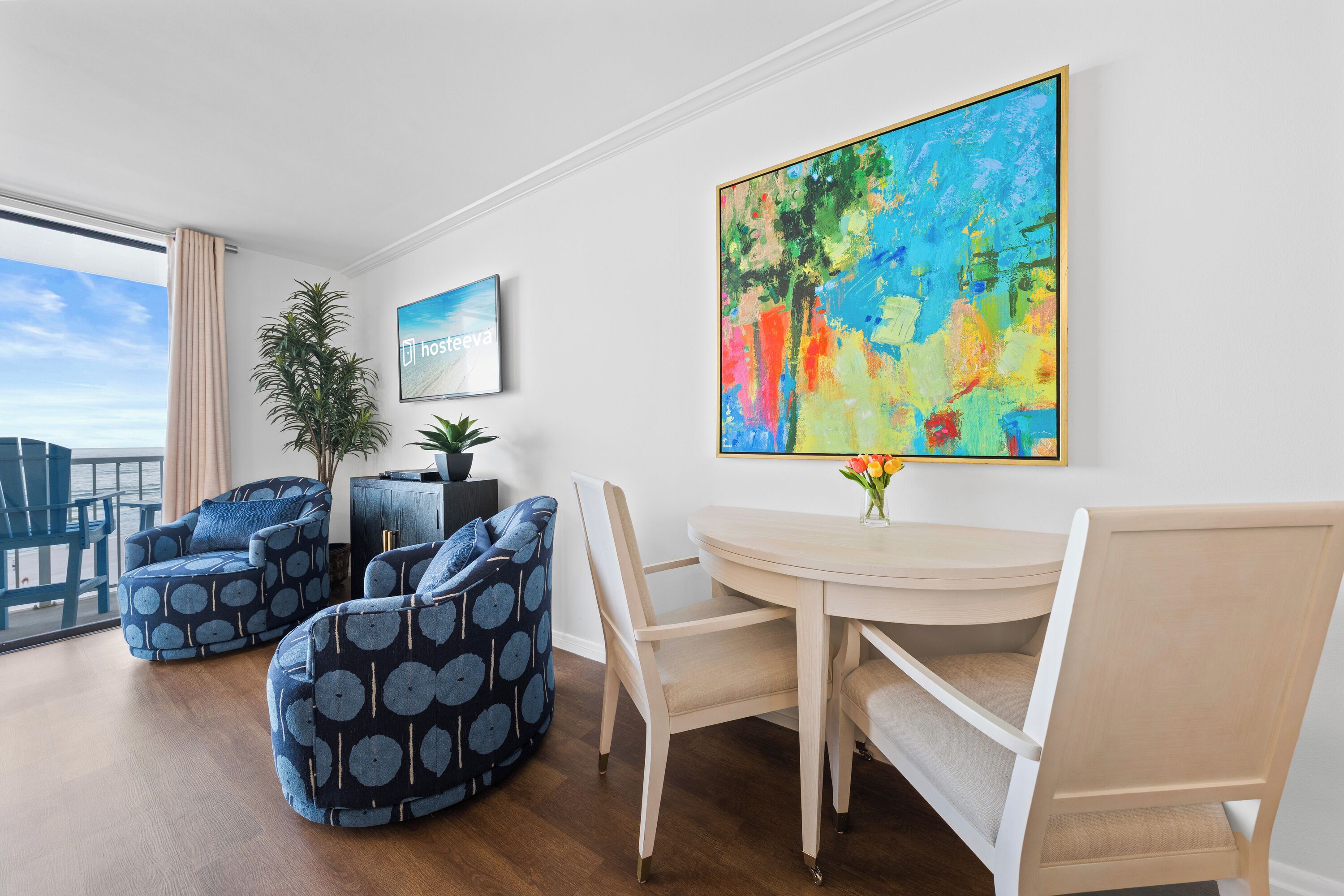 A modern living room with a large window, a dining table, two bean bag chairs, and a colorful abstract painting on the wall.