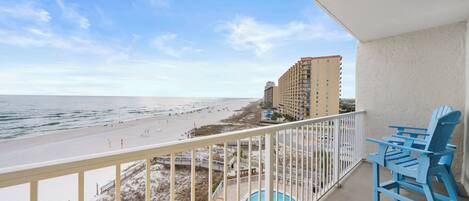 A balcony with a view of the beach and ocean, a pool, and blue and yellow chairs.