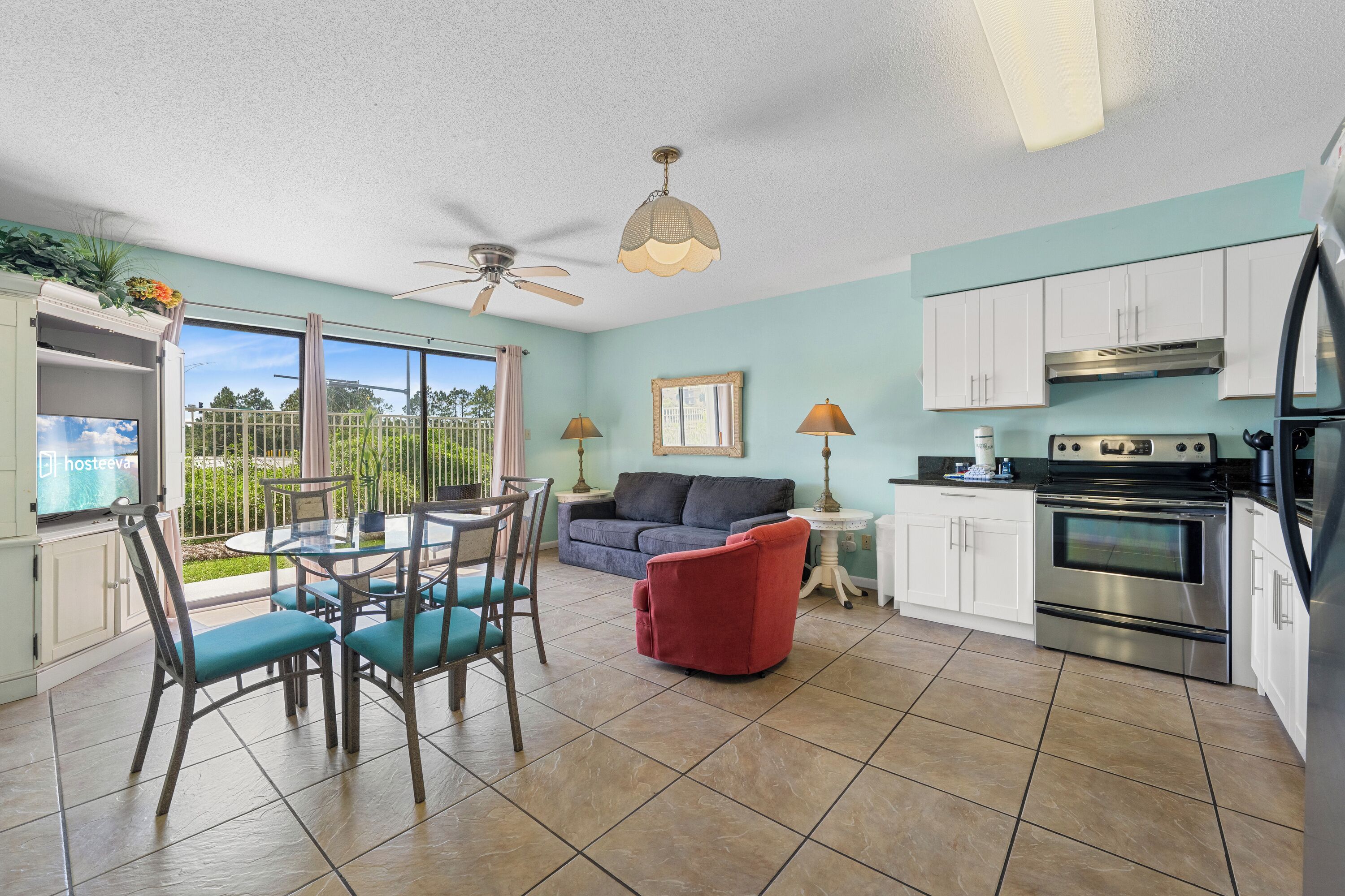 A modern kitchen with white cabinets, a red chair, a dining table with chairs, a ceiling fan, and a large window with a view of greenery.