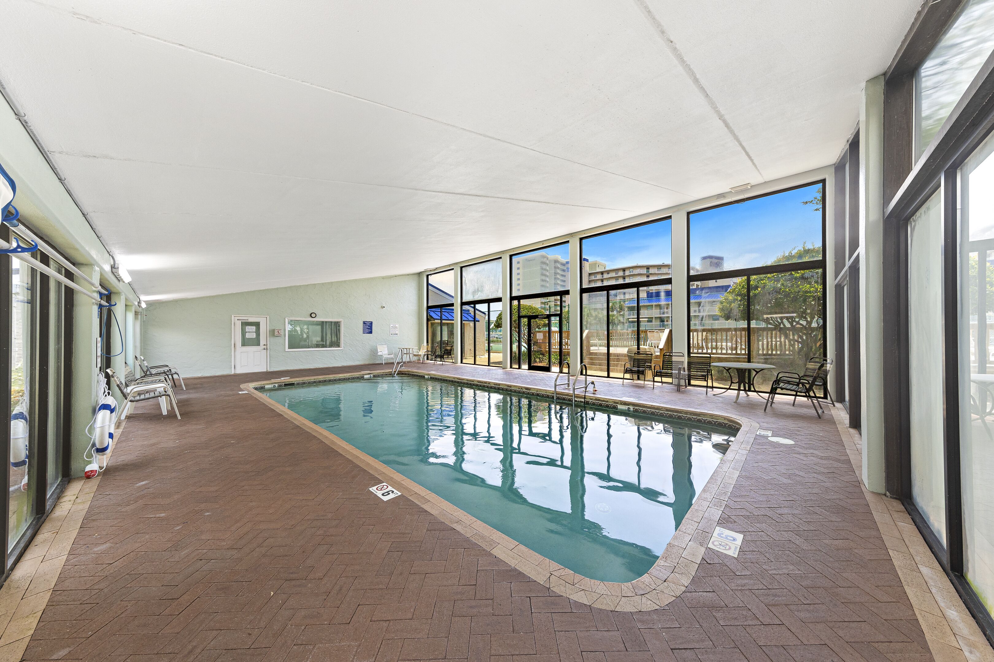 An indoor swimming pool with clear glass walls, a tiled floor, and a few lounge chairs.