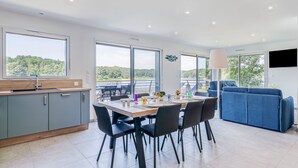 A modern kitchen with a dining area, large windows, and a blue sofa. - Les Roseaux - Vue sur le lac avec jacuzzi (Sanchey)