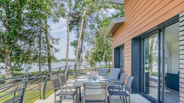 An outdoor dining area with a table set for four, overlooking a body of water, with a modern building and trees in the background. - Les Roseaux - Vue sur le lac avec jacuzzi (Sanchey)