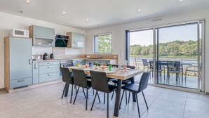 A modern kitchen with a dining area, large windows, and a view of a lake. - Les Roseaux - Vue sur le lac avec jacuzzi (Sanchey)