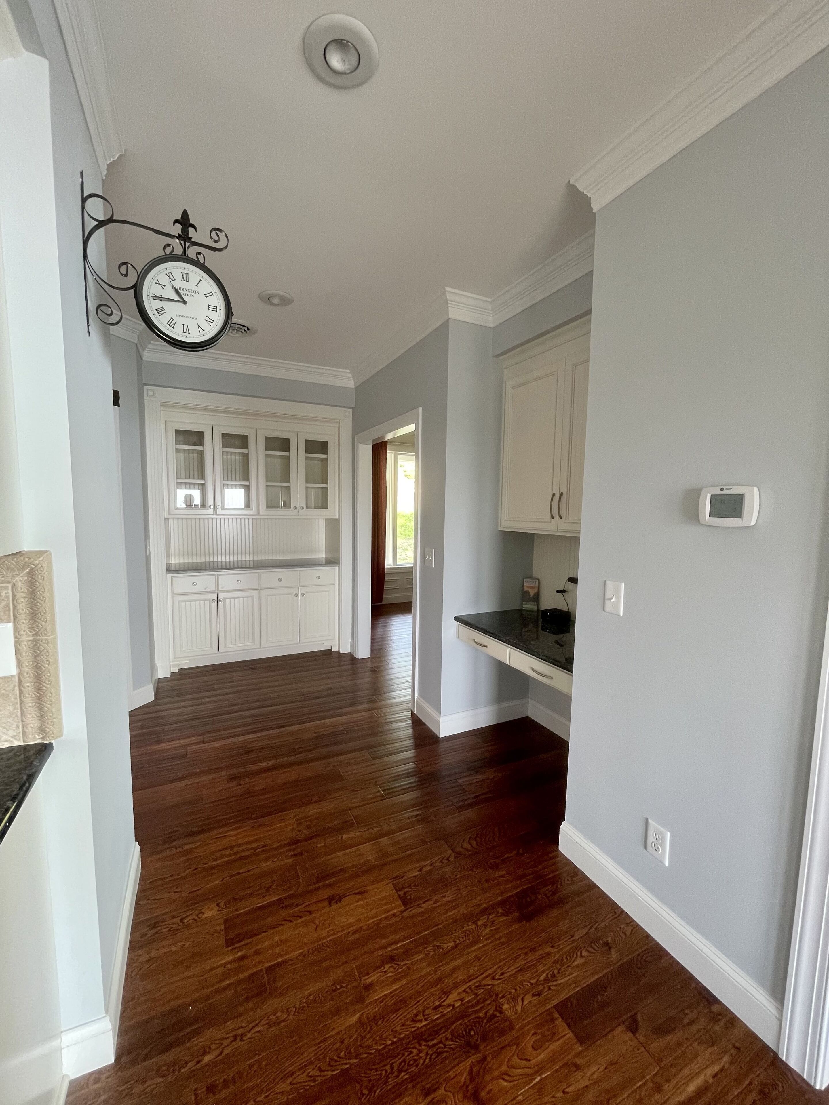 A spacious kitchen with wooden flooring, white cabinetry, and a wall-mounted clock.