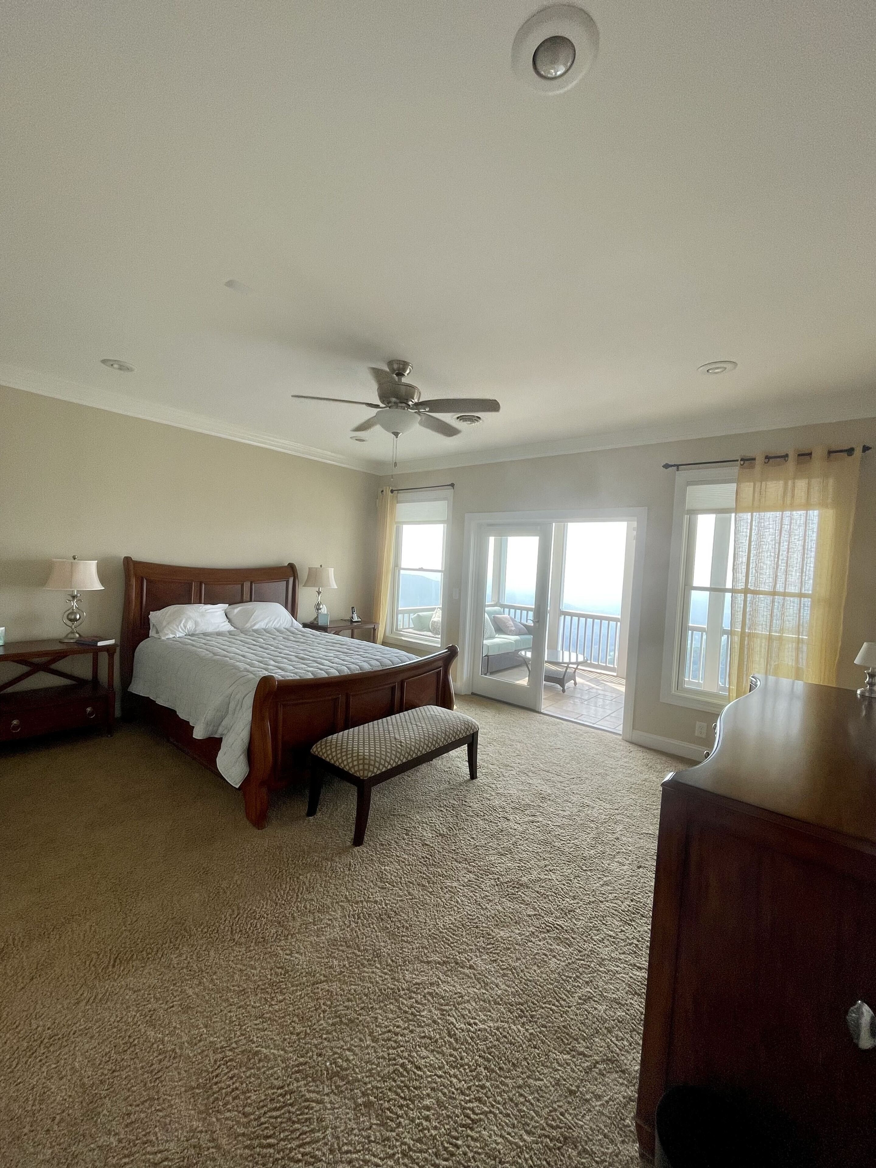 Quarto com cama grande, ventilador de teto e vista para o exterior pelas janelas.