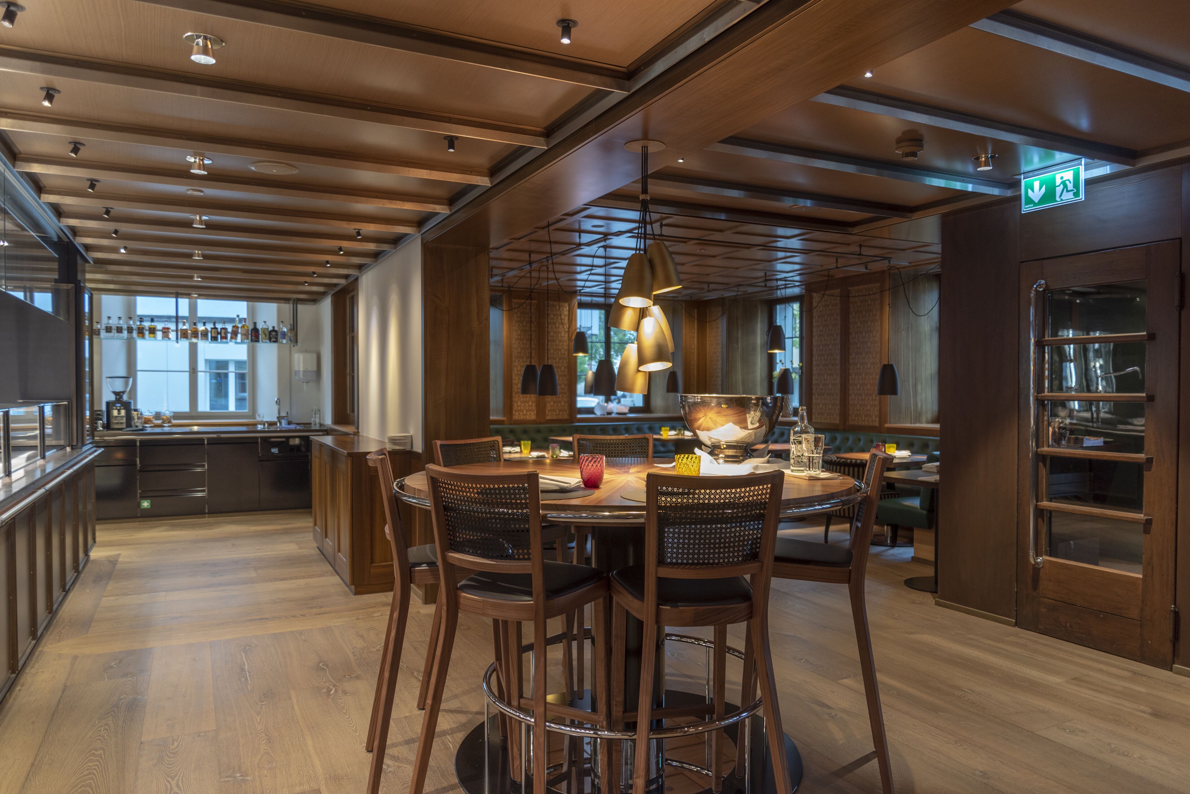 A modern restaurant with a wooden interior, a central dining table with chairs, pendant lighting, and a view of the city through large windows.