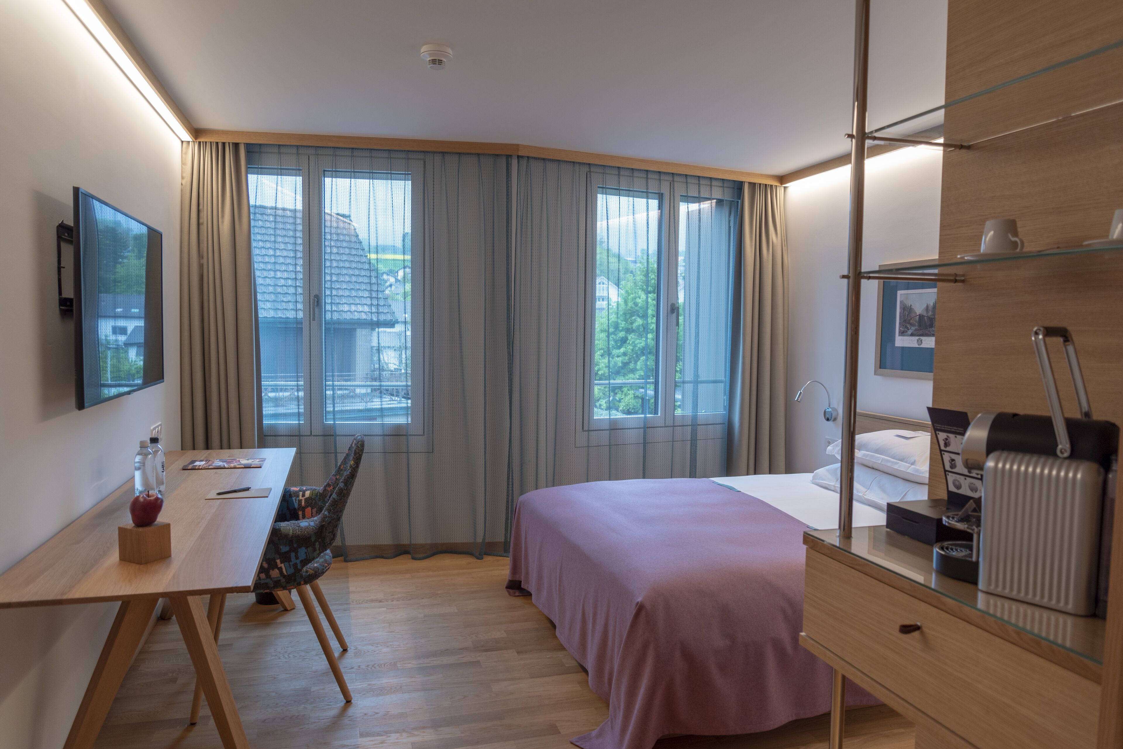 A modern hotel room with a bed, a desk, a chair, and a large window with curtains.