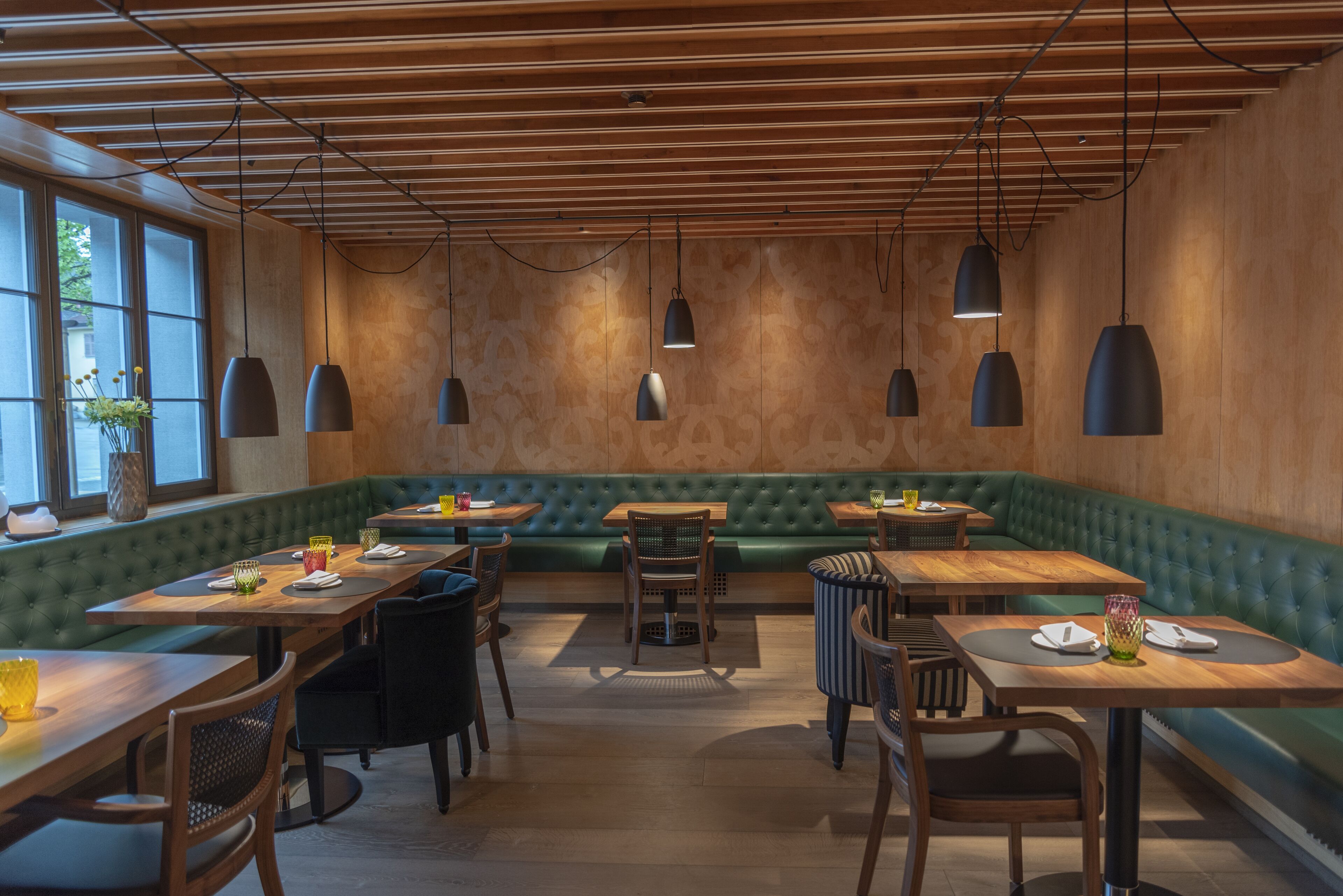 Un restaurante con vigas de madera en el techo, asientos acolchados verdes e iluminación colgante. Las mesas están dispuestas con platos y vasos.