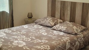 A bed with a floral-patterned bedspread and two pillows, a round side table with a lamp, and a wooden headboard. - La Belle Auvergne 1 / F2 - 3 people (Condat)
