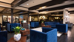 A lobby with blue upholstered seating, a coffee table, and a plant. There is a store in the background and a reception area with a chandelier.