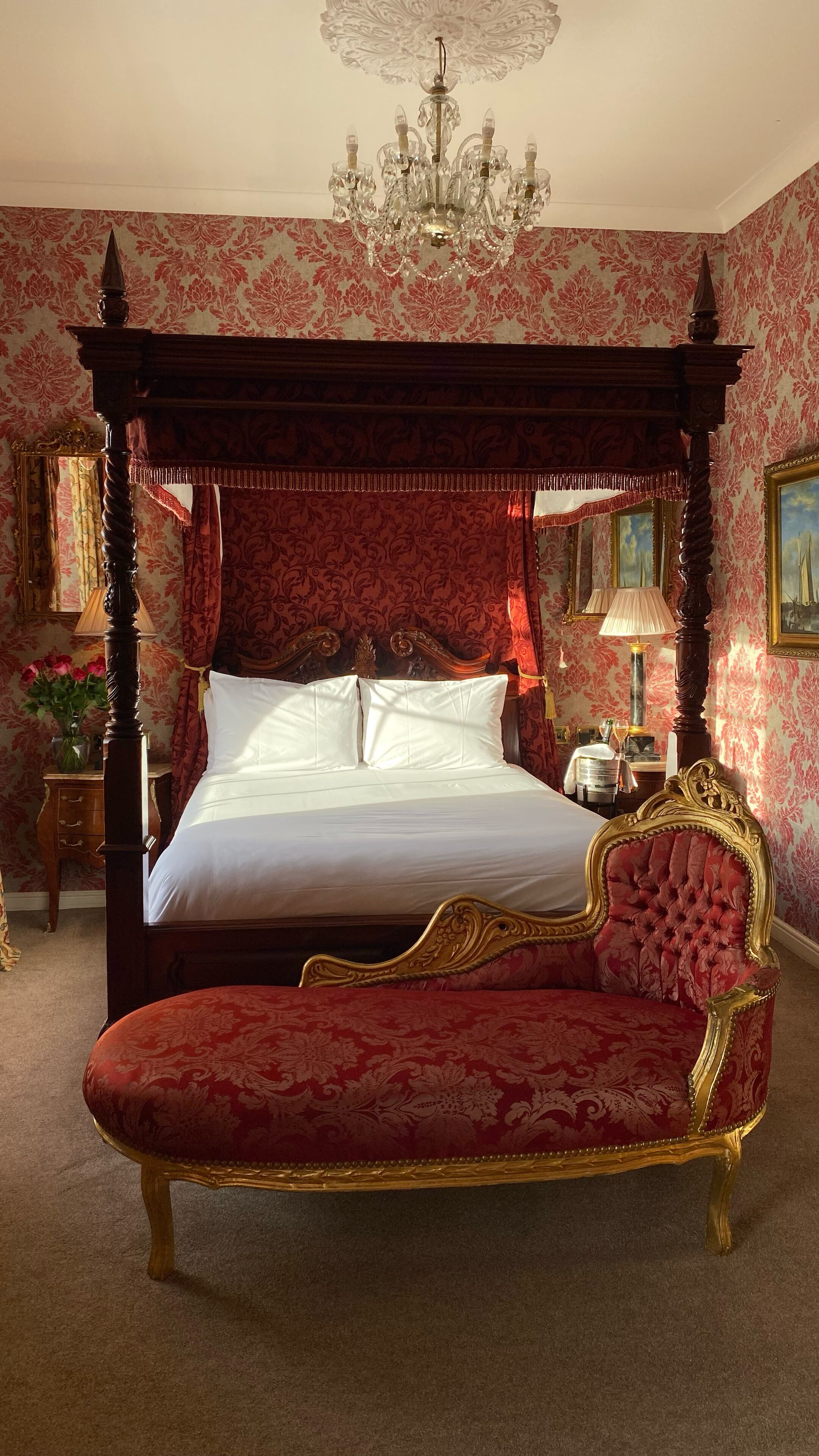A four-poster canopy bed with a red upholstered bench in front, a chandelier, and ornate wallpaper.