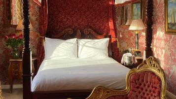 A four-poster canopy bed with a red upholstered bench in front, a chandelier, and ornate wallpaper.