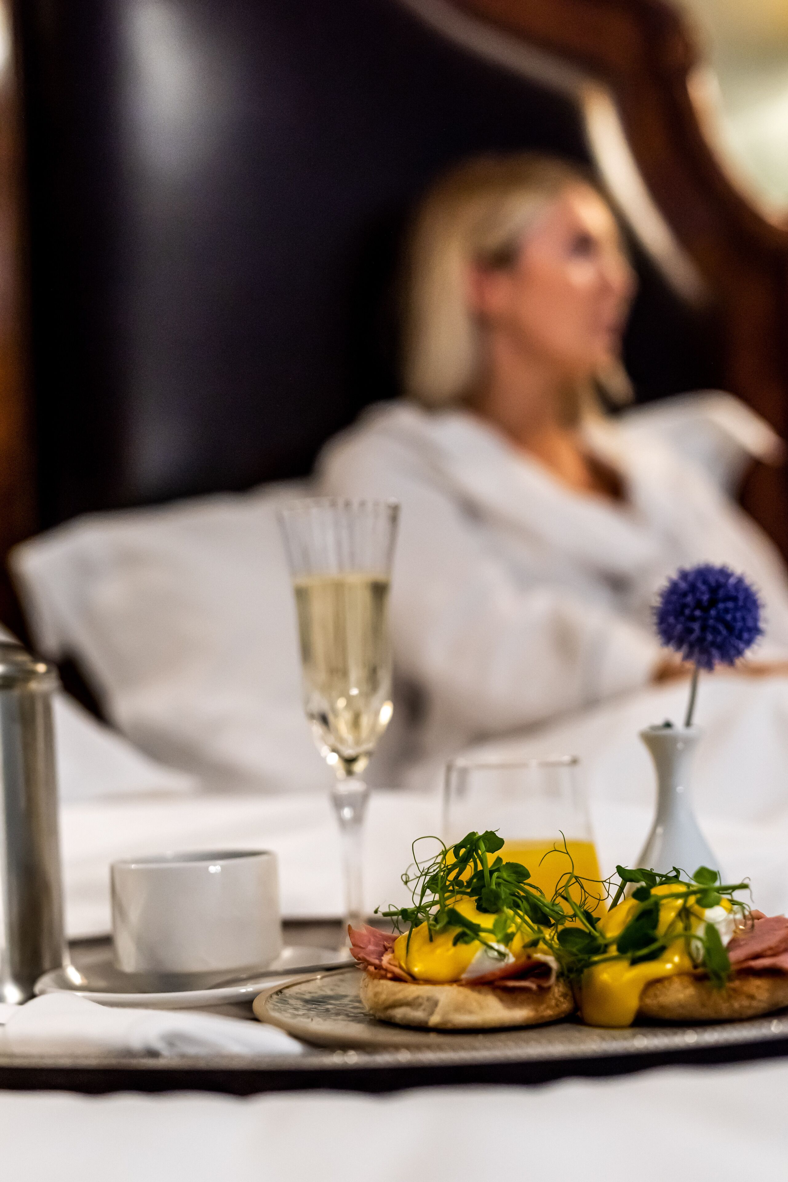 A breakfast tray with eggs benedict, a glass of champagne, and a small vase with a blue flower.