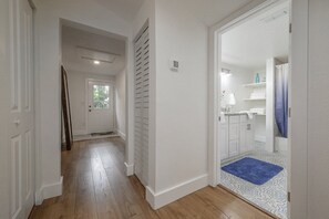 A hallway with wooden flooring, white walls, and a blue rug leading to a bathroom with white cabinets and a shower.