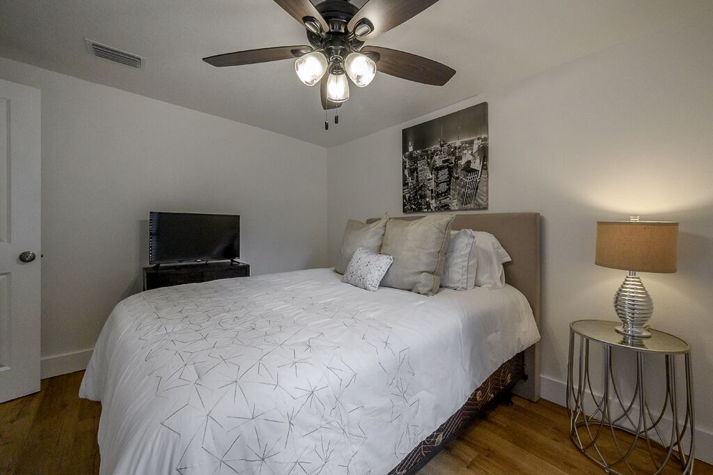 Une chambre à coucher avec un lit, un ventilateur de plafond, une télévision, une lampe et un tableau représentant un paysage urbain accroché au mur.