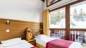 Habitación de hotel con dos camas, cabecero de madera, un oso de peluche en una cama y una ventana con vistas a un paisaje nevado.