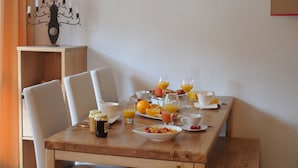 En matplats med ett träbord dukat för frukost, med tallrikar med flingor, frukt och glas med juice. Runt bordet står vita stolar, och på väggen hänger en inramad bild av ett snötäckt landskap.