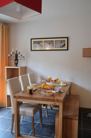 A dining area with a wooden table set for breakfast, including plates of cereal, fruit, and juice glasses. There are white chairs around the table, and a framed picture of a snowy landscape hangs on the wall.