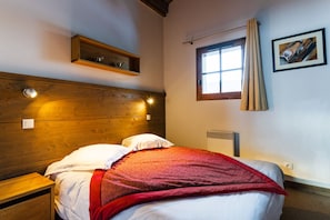 A bedroom with a wooden headboard, a window with curtains, a bedside table, and a wall-mounted shelf.