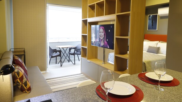 Een hotelkamer met een televisie, een eettafel met borden en glazen, een bank met een gekleurd kussen, en een balkon met stoelen.