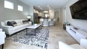 Ein modernes Wohnzimmer mit einem weißen Sofa, einem Couchtisch und einem Fernseher auf einem Standfuß. Im Hintergrund befindet sich eine Küchenzeile mit einem Kühlschrank und einer Theke.