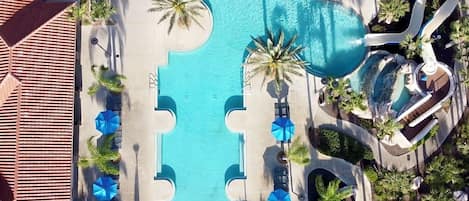 An aerial view of a resort with a central swimming pool, surrounded by palm trees and lounge chairs.