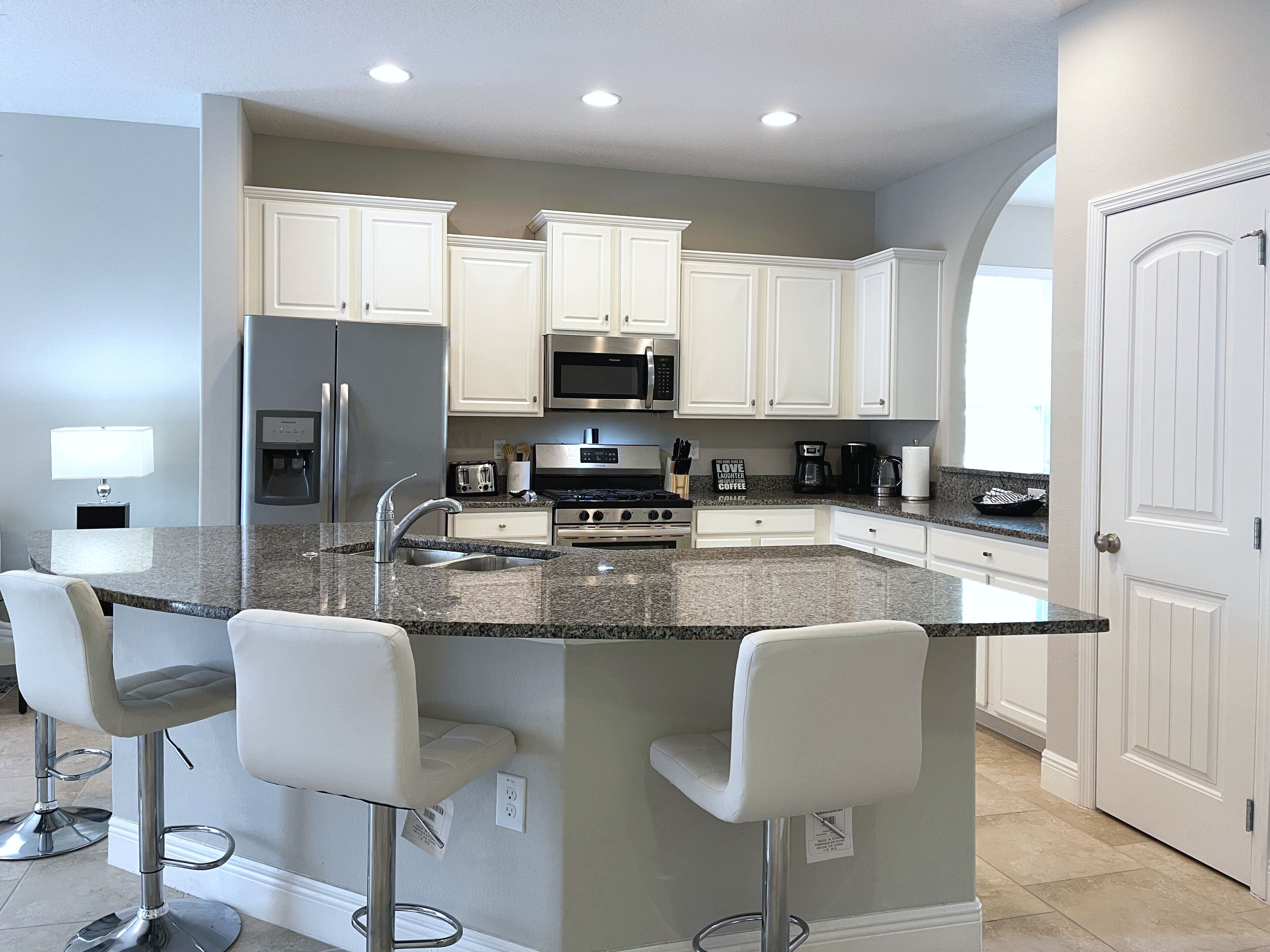 A modern kitchen with white cabinets, granite countertops, stainless steel appliances, and a bar area with white stools.