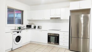 Cozinha moderna com armários brancos, geladeira de aço inoxidável, máquina de lavar, micro-ondas, torradeira e forno compacto.