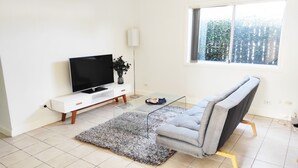 Uma sala de estar moderna com uma televisão de tela plana sobre um console branco, um sofá cinza, uma mesa de centro de vidro e uma janela grande com vista para vegetação.