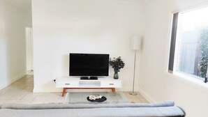 Uma sala de estar moderna com uma televisão de tela plana sobre um console branco, um vaso com flores escuras, um abajur e um sofá cinza.