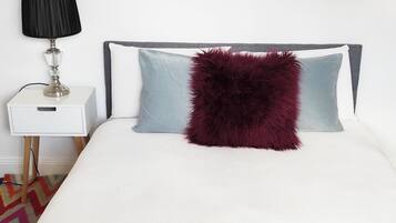 A neatly made bed with a white comforter, a dark red fluffy pillow, and two folded towels in teal and gray. A white bedside table with a black lamp is to the left.