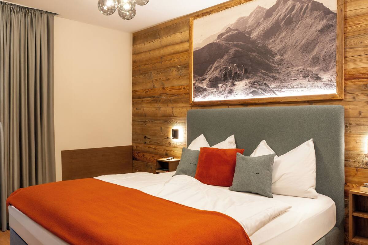 Una habitación de hotel con cabecera de madera, un cuadro grande con paisaje de montañas, una cama con ropa de cama blanca y naranja, y una mesita de noche con lámpara.