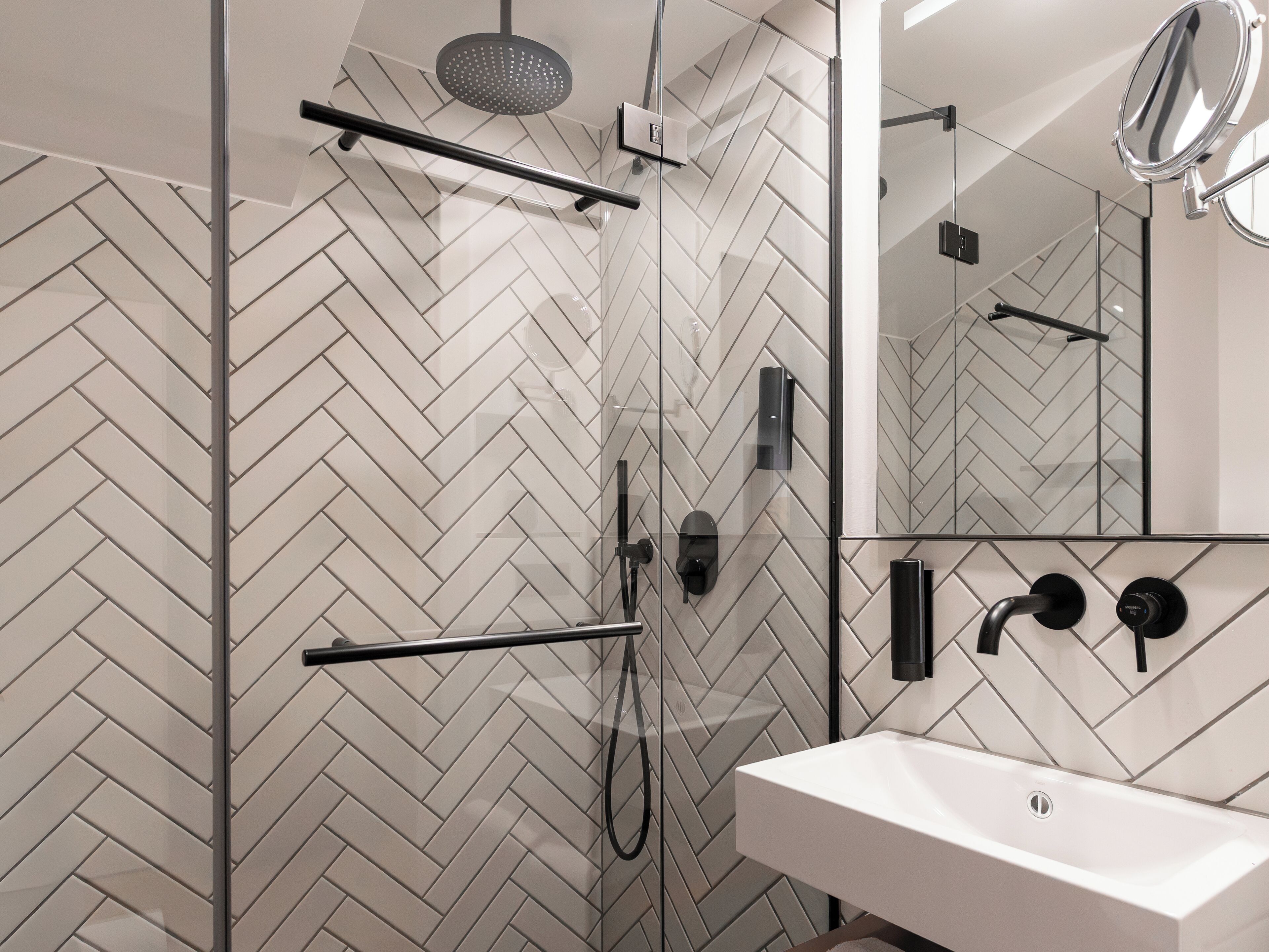 A modern bathroom with a herringbone tile backsplash, a glass shower enclosure, a white sink, and a mirror.