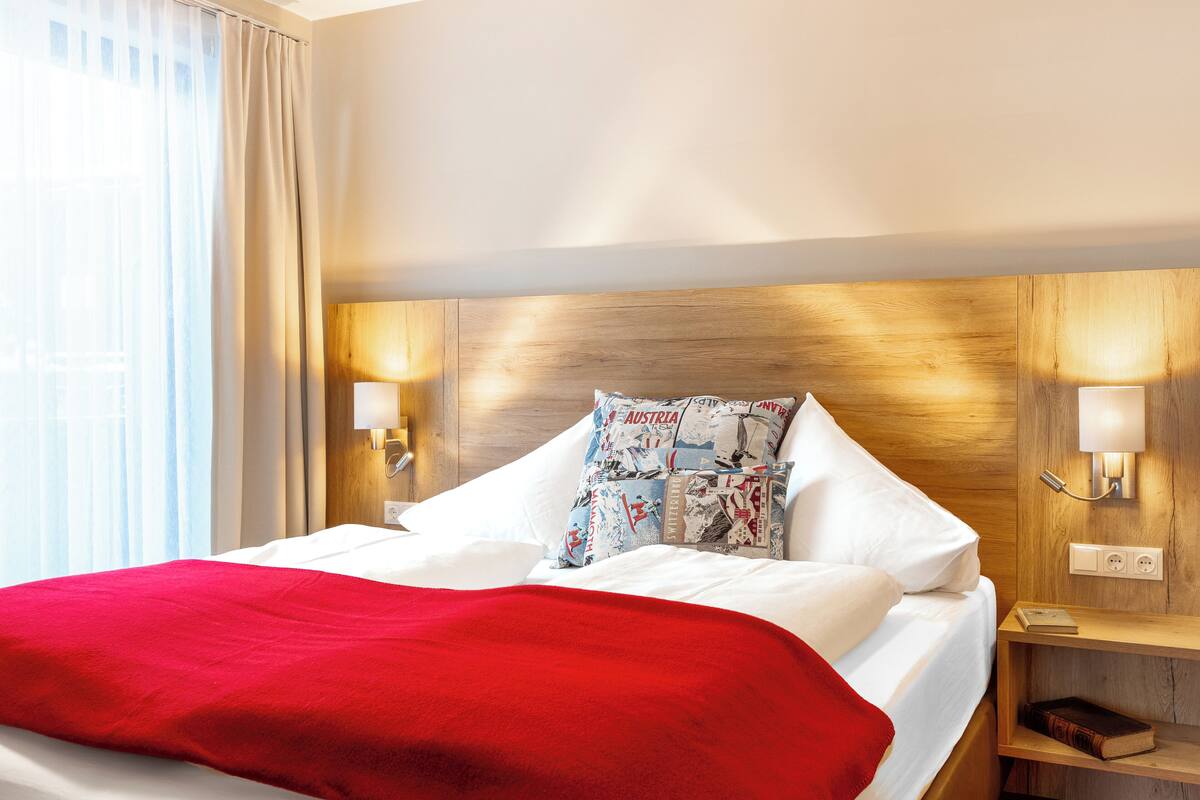Una habitación de hotel con cabecera de madera, ropa de cama blanca, una manta roja y una mesita de noche con una lámpara y un libro.