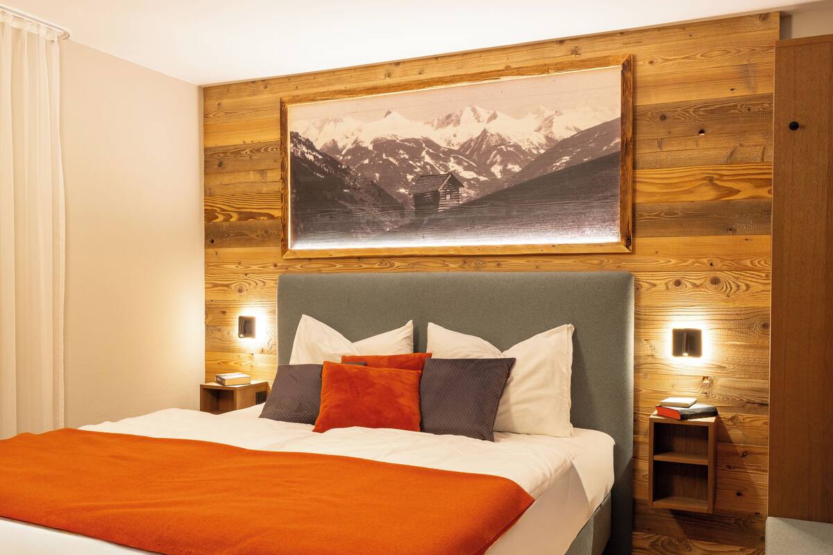 Habitación de hotel con una cama grande, paneles de madera en las paredes, un cuadro de un paisaje de montaña y dos mesitas de noche con lámparas.