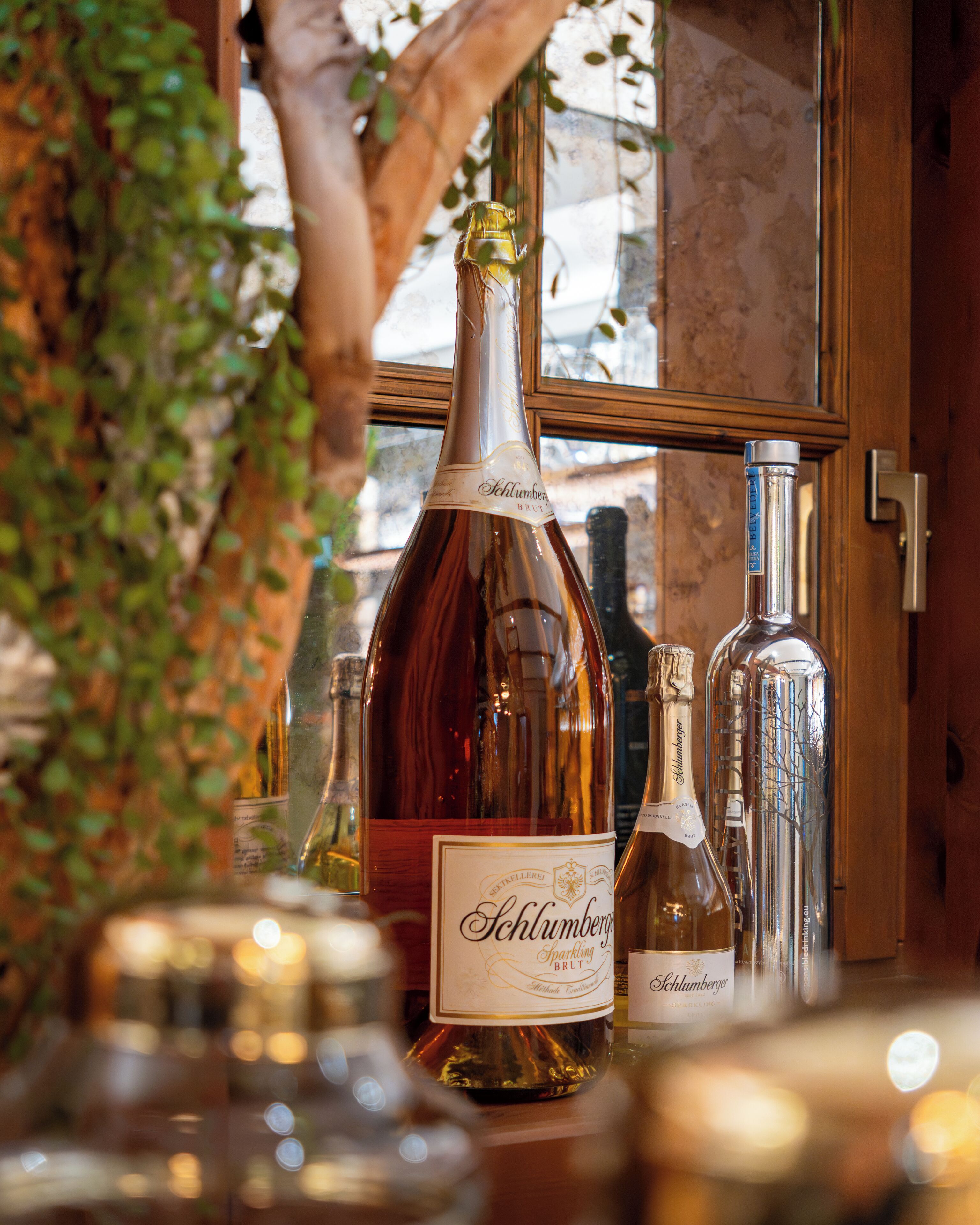 A bottle of Schlumberger sparkling wine prominently displayed on a table with other bottles, set against a wooden backdrop and a window with a view.