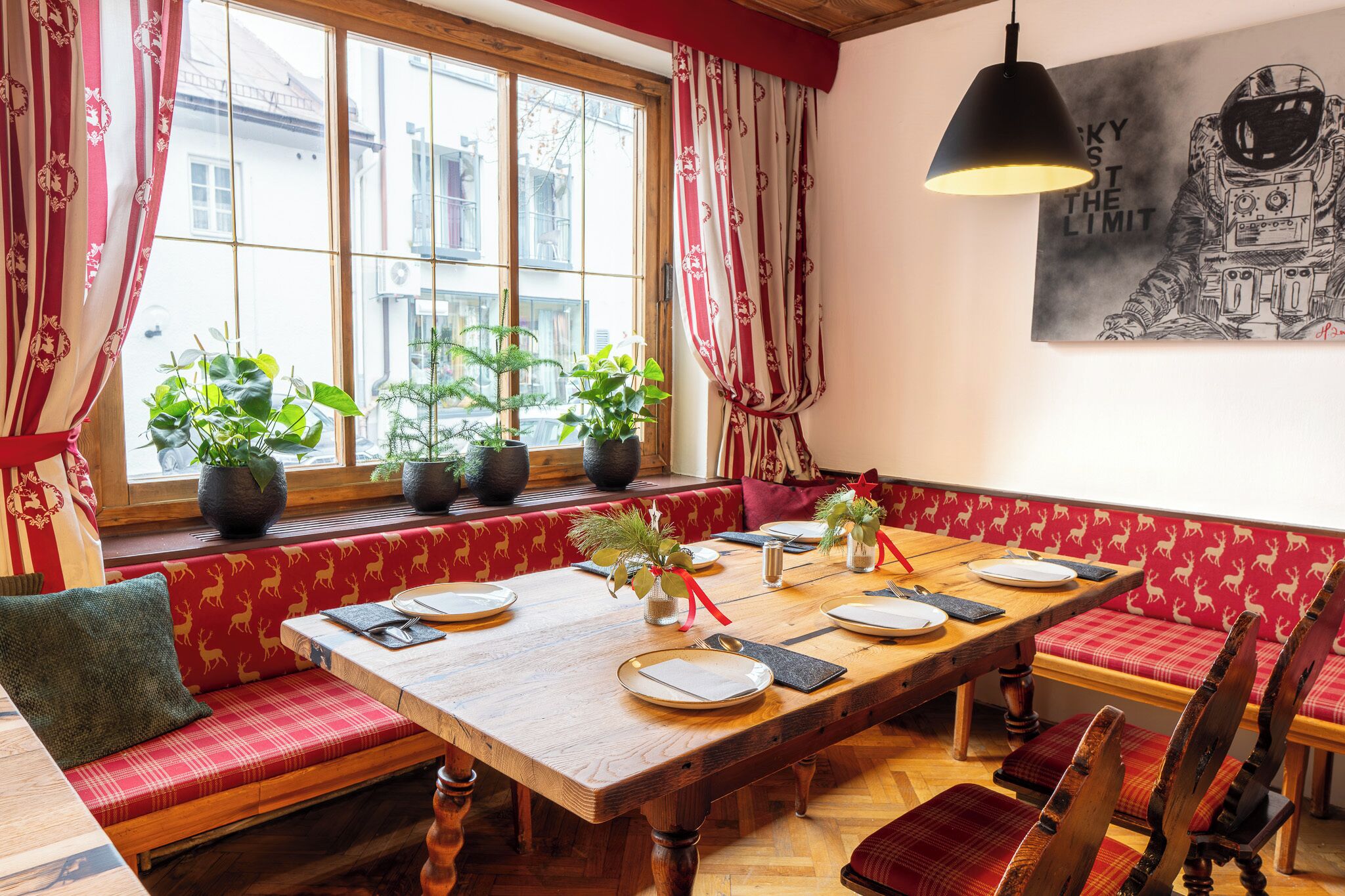 Un coin repas avec une table en bois dressée pour quatre personnes, des bancs à motifs rouges, des plantes en pot et une grande fenêtre avec des rideaux.