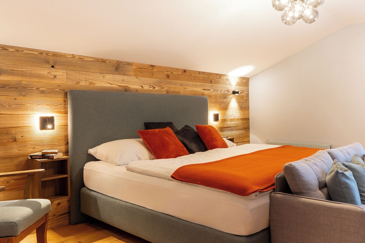 Un dormitorio moderno con cabecera de madera, cama con ropa de cama blanca y detalles naranja, un sillón gris y una mesita de noche de madera con libros.