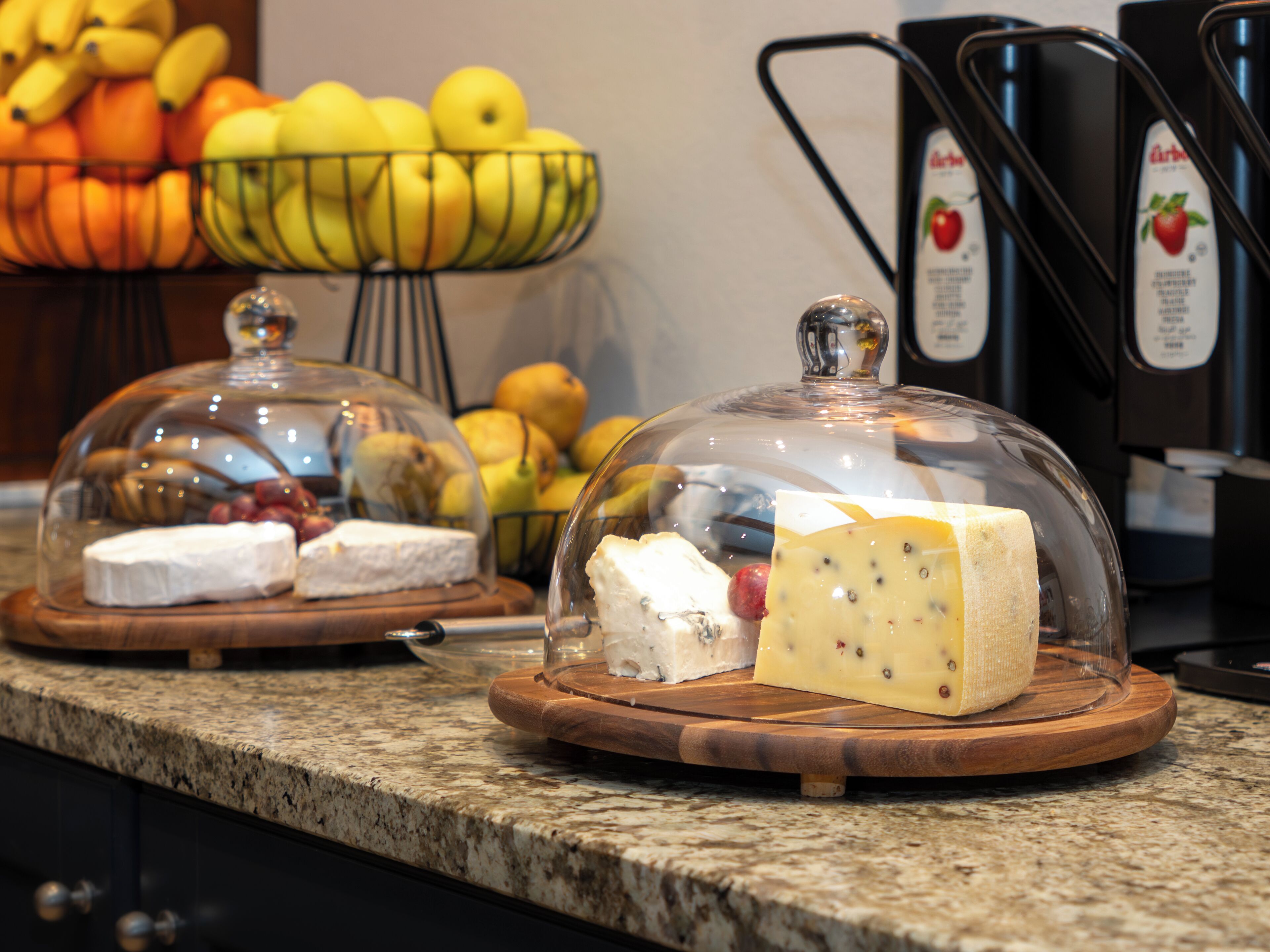 A cheese and fruit display with a glass dome cover on a granite countertop.