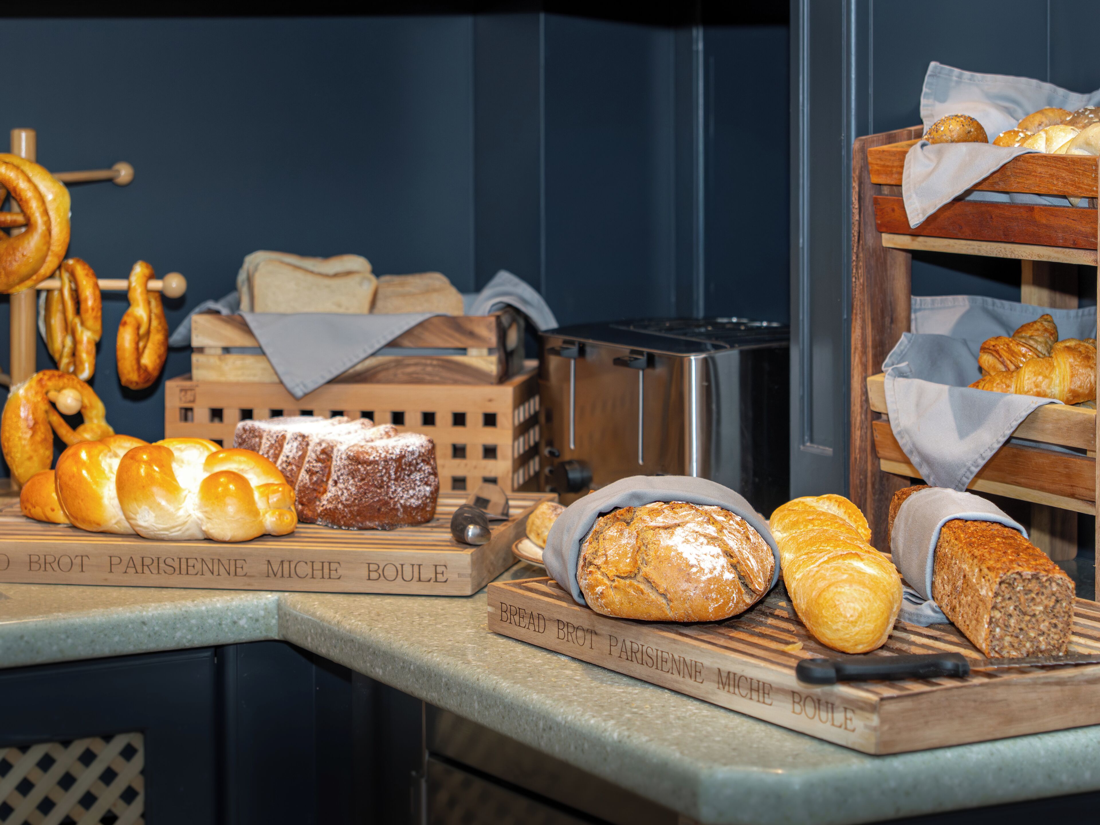 Une variété de pains et de viennoiseries présentés sur des planches en bois, avec une planche à découper, un grille-pain et une boîte à pain en arrière-plan.