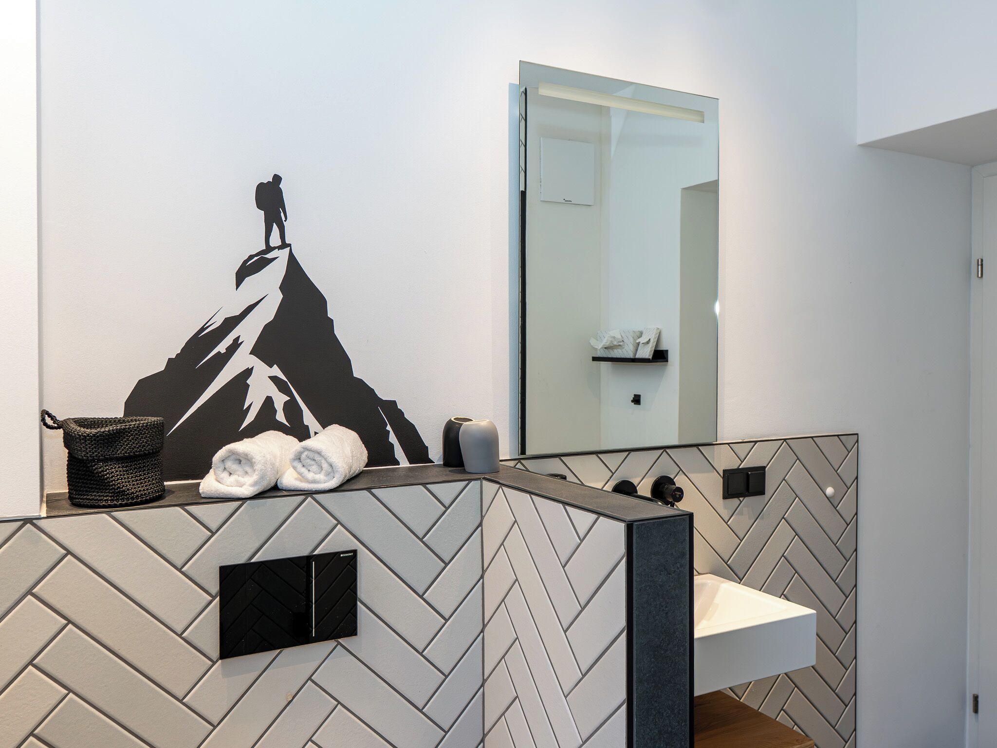 Un baño moderno con salpicadero de baldosas en patrón herringbone, un espejo grande y un adhesivo mural con forma de montaña.