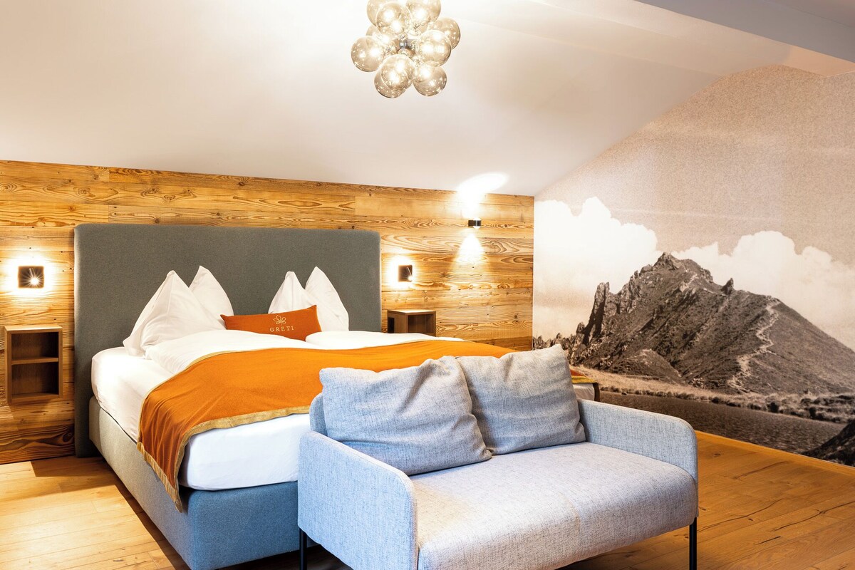 Een moderne slaapkamer met een houten wand, een bed met wit en oranje beddengoed, een grijs gestoffeerde fauteuil en een muurschildering van een berglandschap.