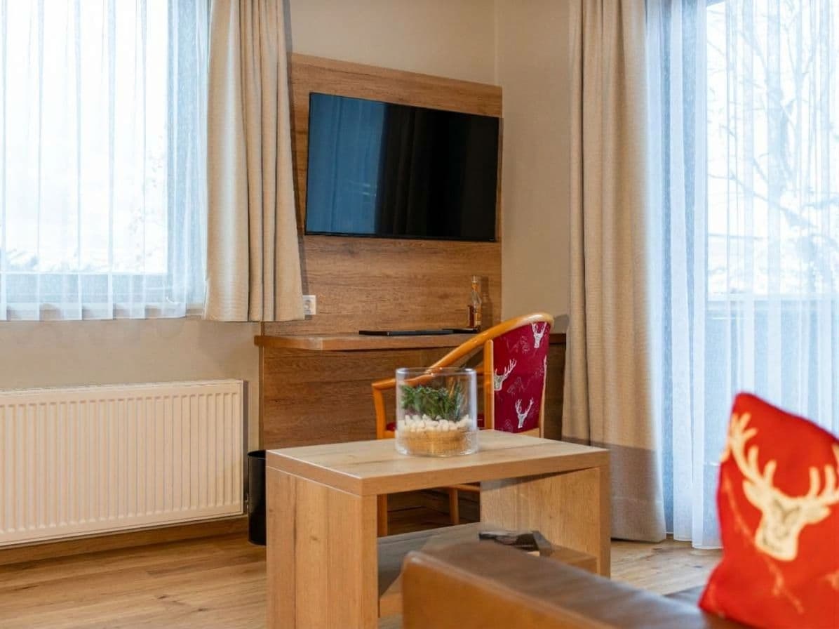 Una habitación con un televisor montado en una pared de madera, una pequeña mesa con un objeto decorativo, una silla roja con estampado de ciervo y un sofá con un cojín rojo.