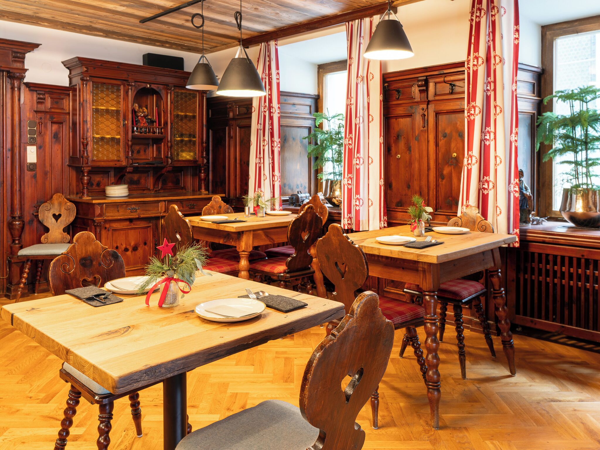 Comedor tradicional con mesas y sillas de madera, un candelabro y cortinas con estampado rojo y blanco.