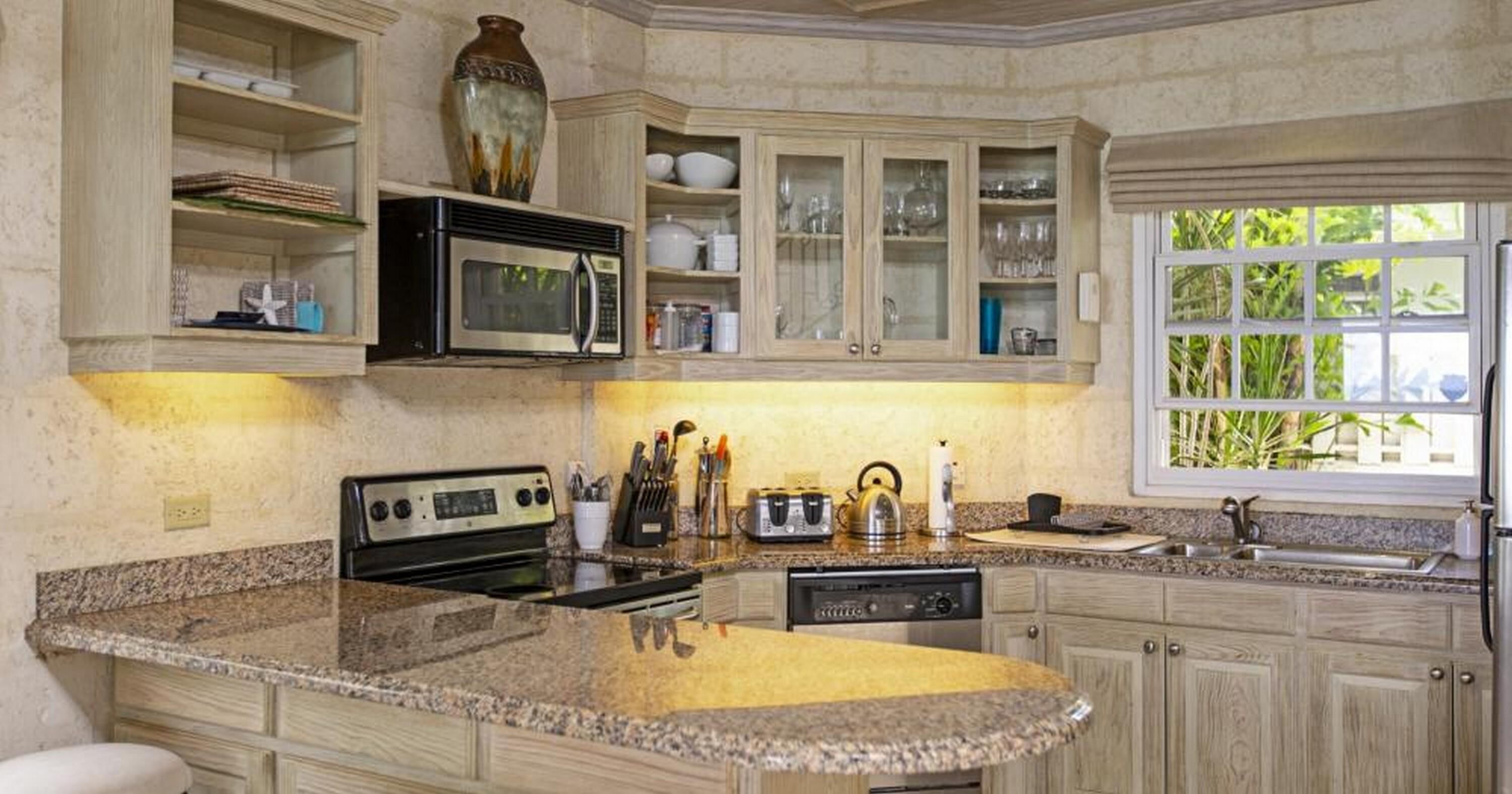 Una cocina con encimeras de granito, estufa, microondas y lavavajillas. Hay armarios con puertas de vidrio y una ventana con vistas a zonas verdes.