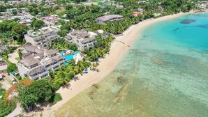 Vista aérea de un complejo turístico costero con varios edificios, una playa y aguas turquesas cristalinas.