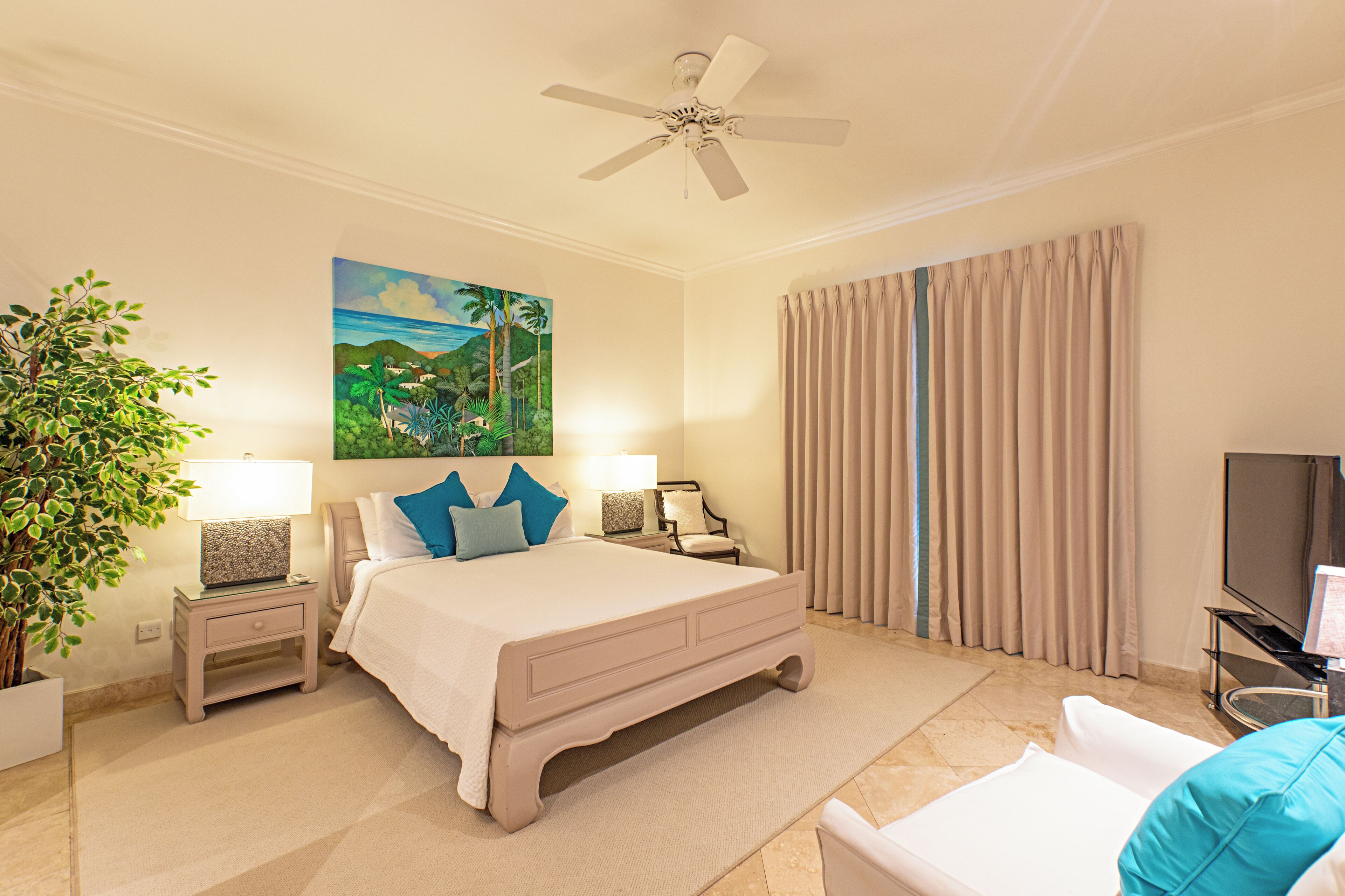 Une chambre d’hôtel avec un grand lit, un ventilateur de plafond, une télévision, une plante et un tableau au mur.