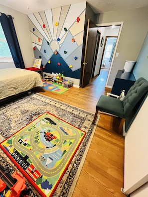 Une salle de jeux pour enfants avec un mur d’escalade, un lit, un tapis de jeu et un ensemble de trains miniatures.