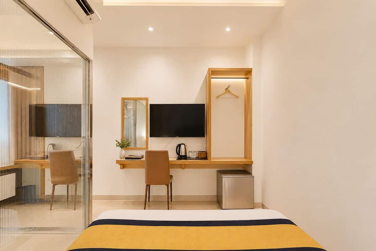 Quarto de hotel moderno com uma cama, uma escrivaninha com televisão, uma cadeira e uma pequena cozinha equipada.